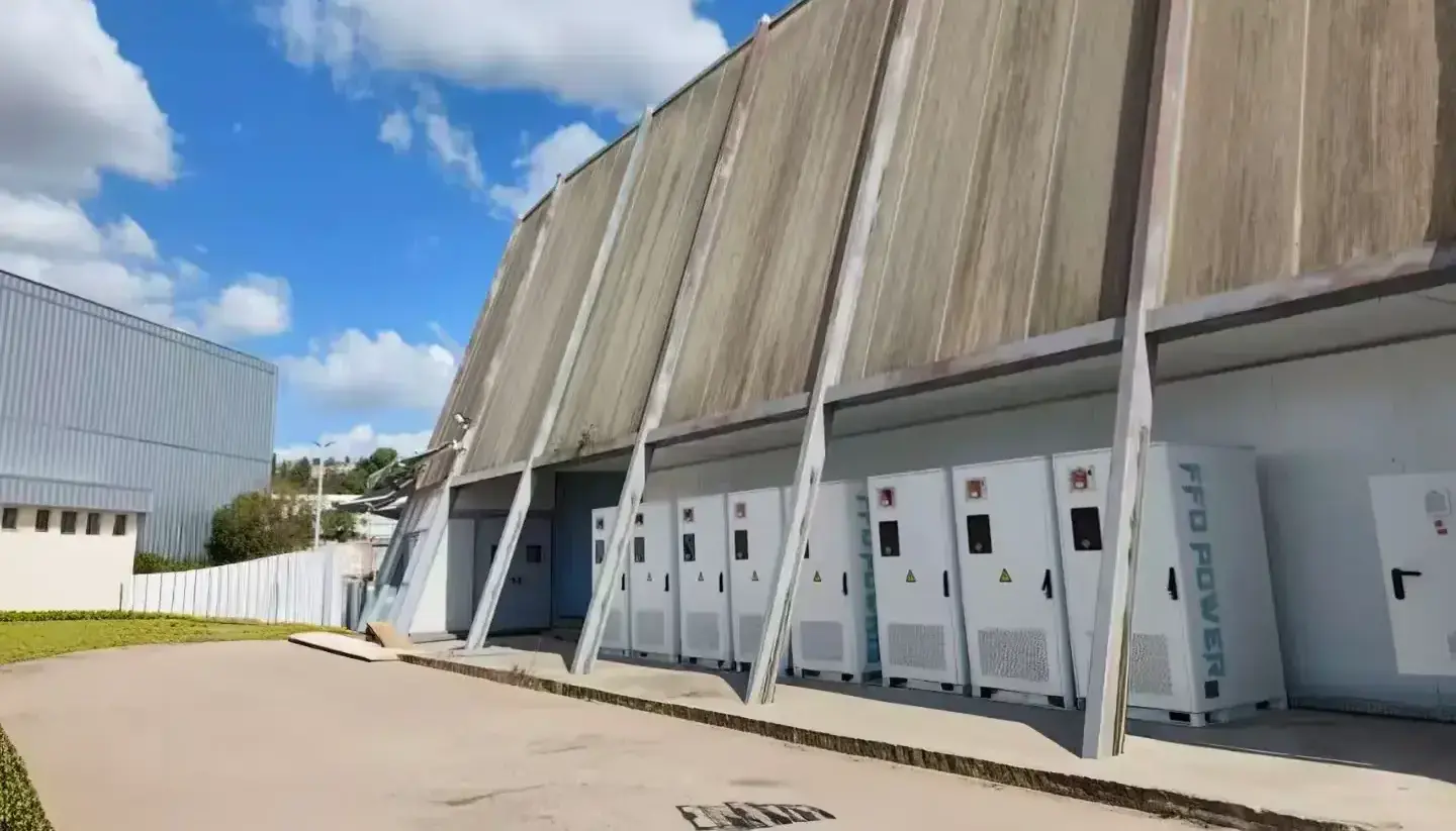 Wide view of the peak-shaving installation showing multiple battery cabinets lined along a long building with a sloped roof.”